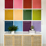 Large undated and reusable wall calendar in colorful rainbow hues from Kaleidoscope Living is hung over a dresser.