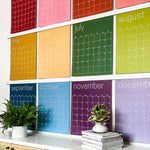 Close up of the large undated and reusable wall calendar in colorful rainbow hues from Kaleidoscope Living hung with custom wood poster hangers.