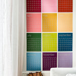 Reusable rainbow wall calendar with twelve individual pages hung on a white wall.
