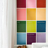 Reusable rainbow wall calendar with twelve individual pages hung on a white wall.