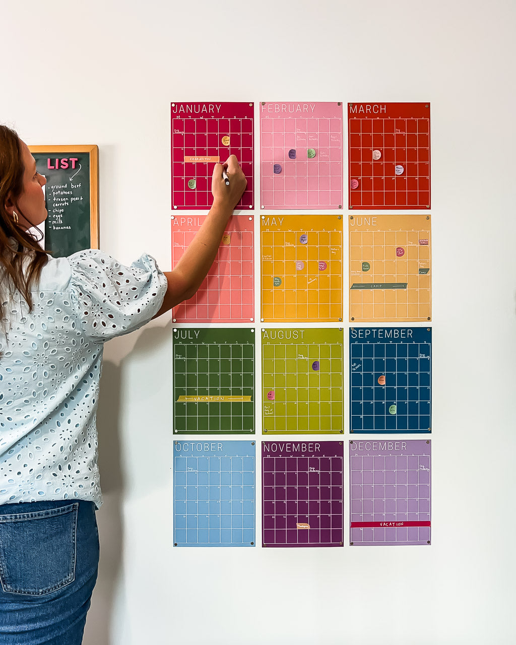 Woman writing on a monthly wall calendar in rainbow colors hanging on a white wall.