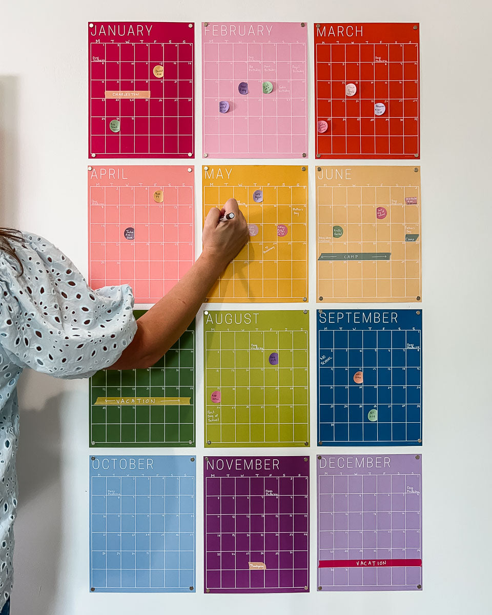 A person is writing on an undated, single-use wall calendars in a rainbow color scheme.
