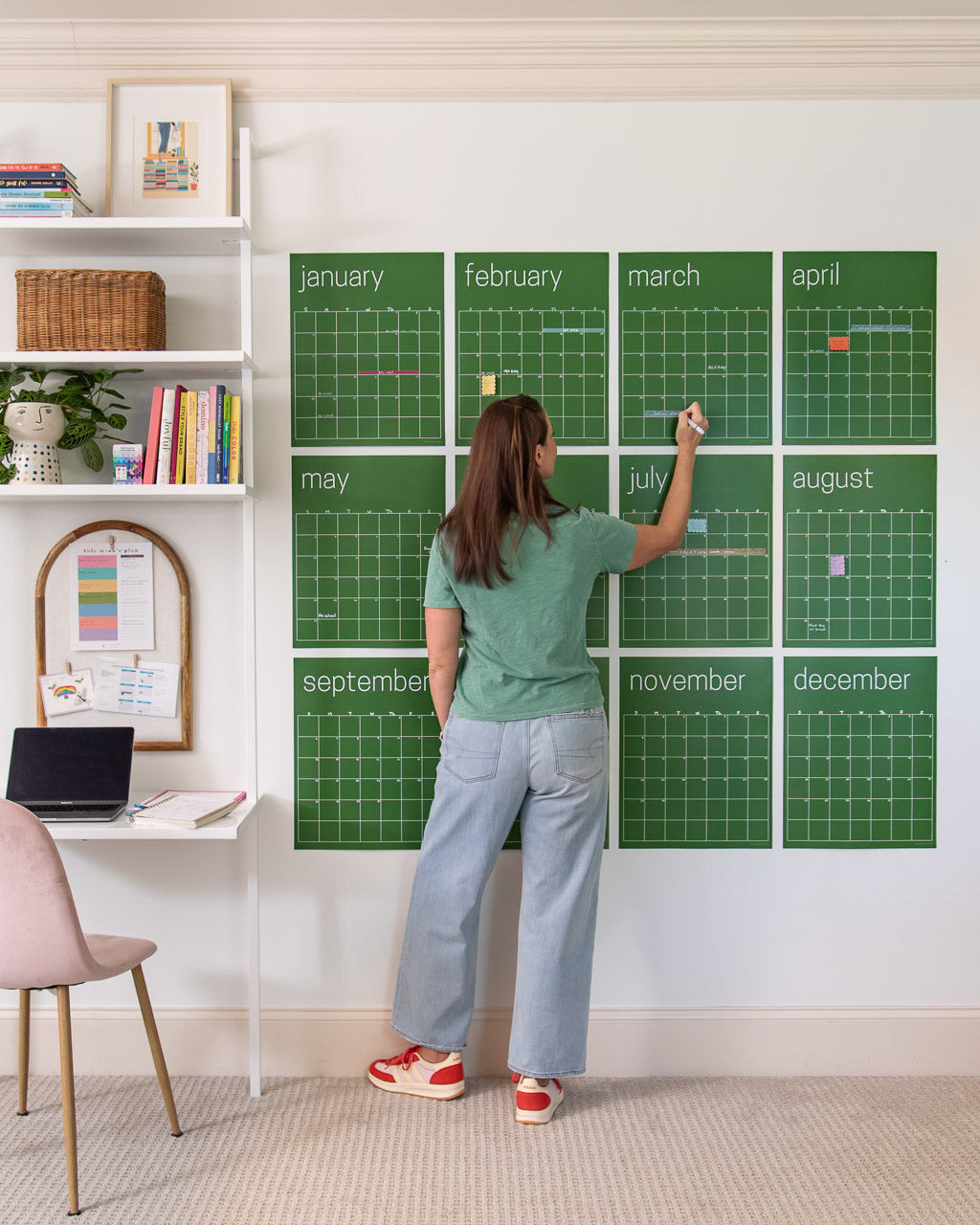 Women writing on a large, undated green wall calendar with twelve monthly pages displaying sticky notes and washi tape.