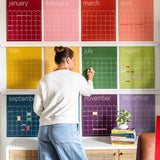 woman writing on large reusable wall calendar in colorful rainbow hues from Kaleidoscope Living