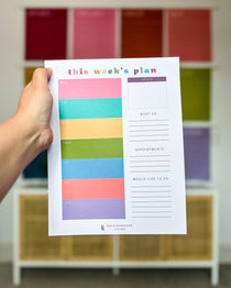 A colorful weekly planning notepad with colorful blocks for each week day being held in front of a large wall calendar by Kaleidoscope Living.