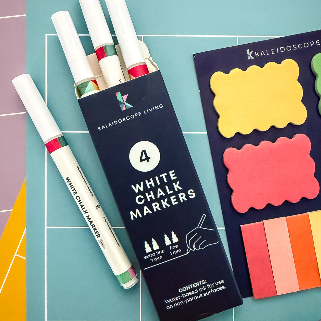 Set of four fine-tip white chalk markers next to colorful sticky notes on a stack of monthly calendar pages